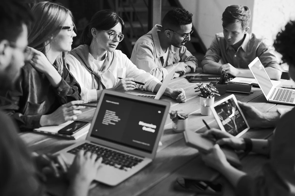Group of six young professionals sitting around a table working on laptops and tablets during a collaborative meeting.
